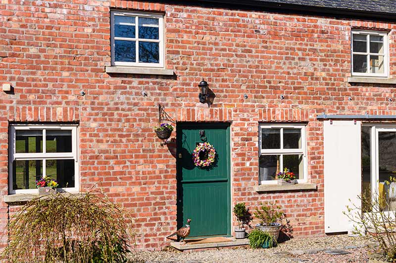 Brick cottage with green door and white-framed windows, adorned with a wreath.